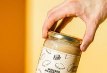 Hand hält ein Glas weißes Mandelmus von Korodrogerie über Brot und Mandeln