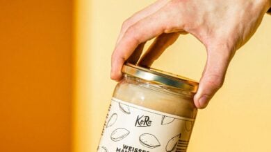 Hand hält ein Glas weißes Mandelmus von Korodrogerie über Brot und Mandeln