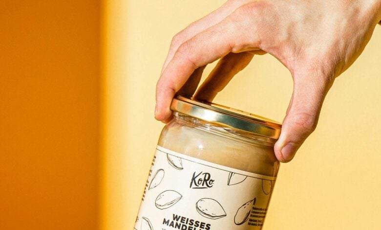 Hand hält ein Glas weißes Mandelmus von Korodrogerie über Brot und Mandeln